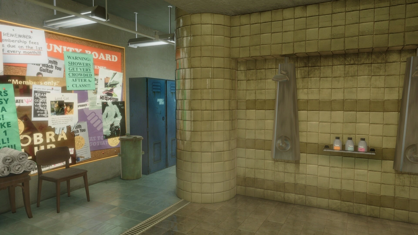 An empty tiled gym shower room contains blue lockers, towels and a crowded noticeboard.